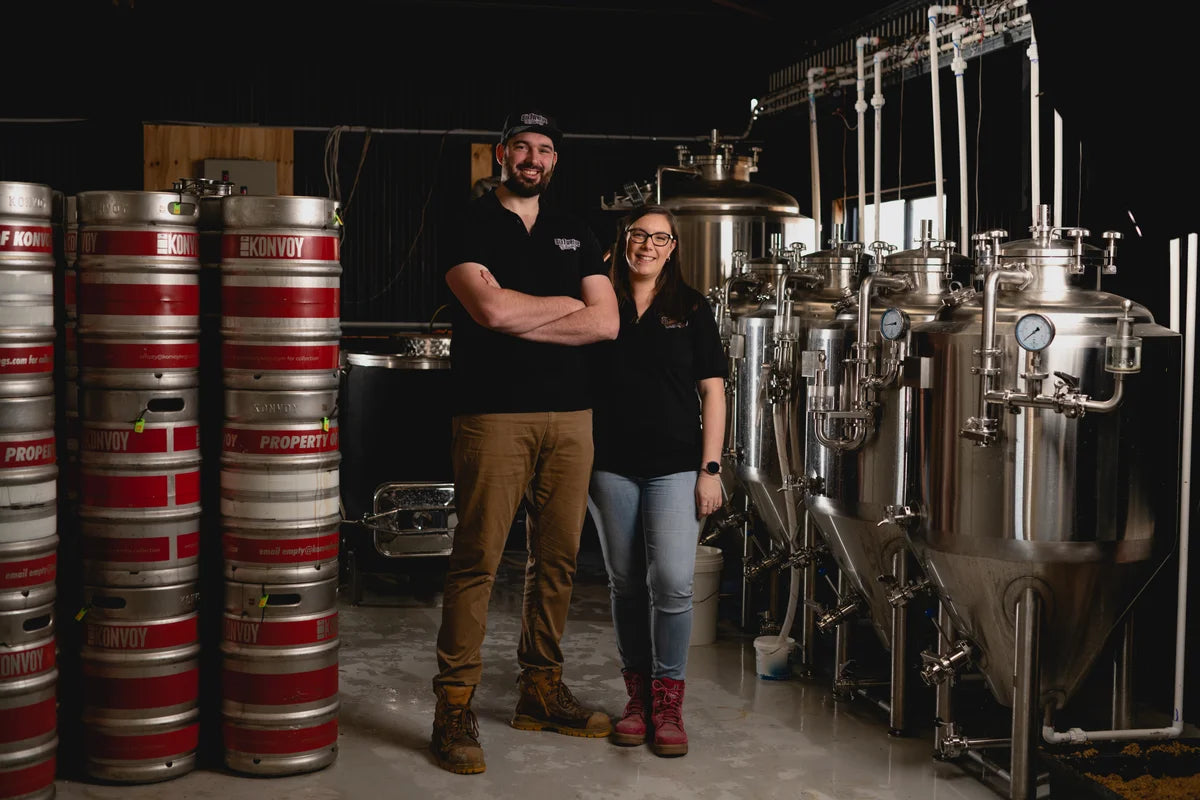 Bradley and Alison Reid standing in brewery with beer kegs to the left and fermenters to the right