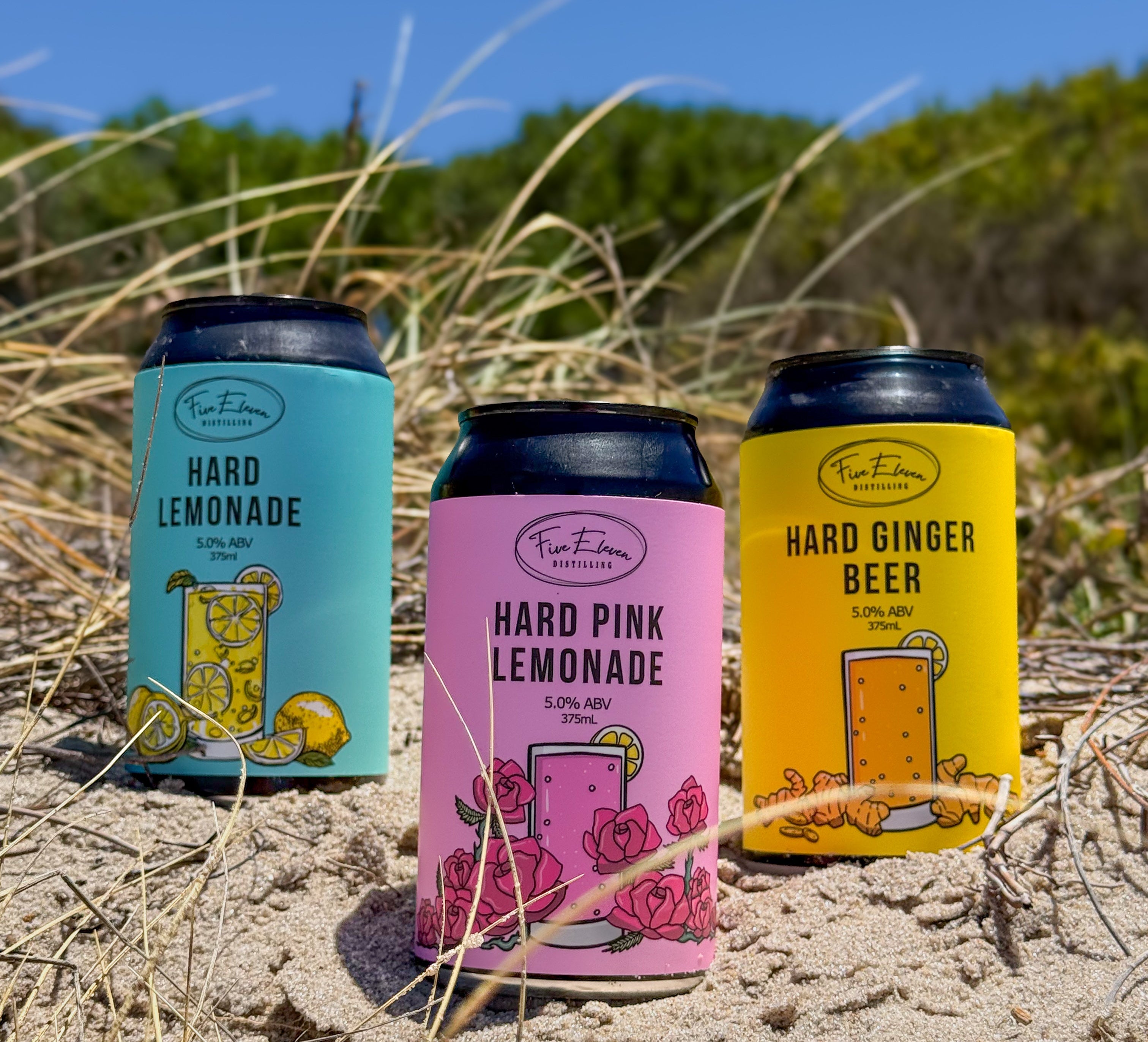 Three colourful cans of Five Eleven Distilling Hard Drinks on a sandy beach with grass and blue sky.